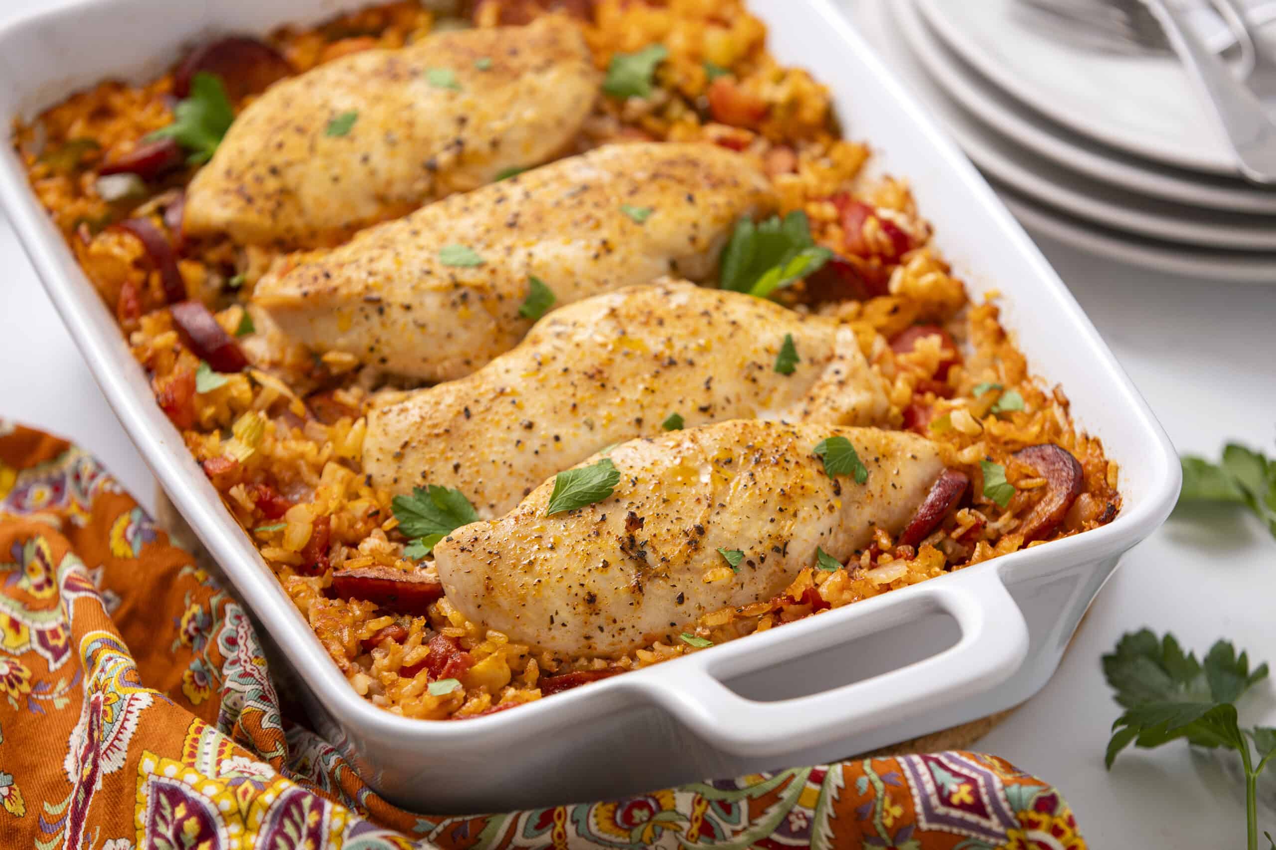Juicy baked chicken breasts with rice and vegetables in a white baking dish. Perfect for a hearty, flavorful dinner recipe. Garnished with fresh parsley, served with stacked white plates.