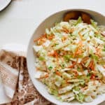 Fresh homemade coleslaw salad with shredded cabbage, carrots, and green onions in a white bowl on a light wooden table.