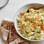 Fresh homemade coleslaw salad with cabbage, carrots, and green onions in a white ceramic bowl on a rustic white wooden table. Perfect for summer picnics and barbecues.