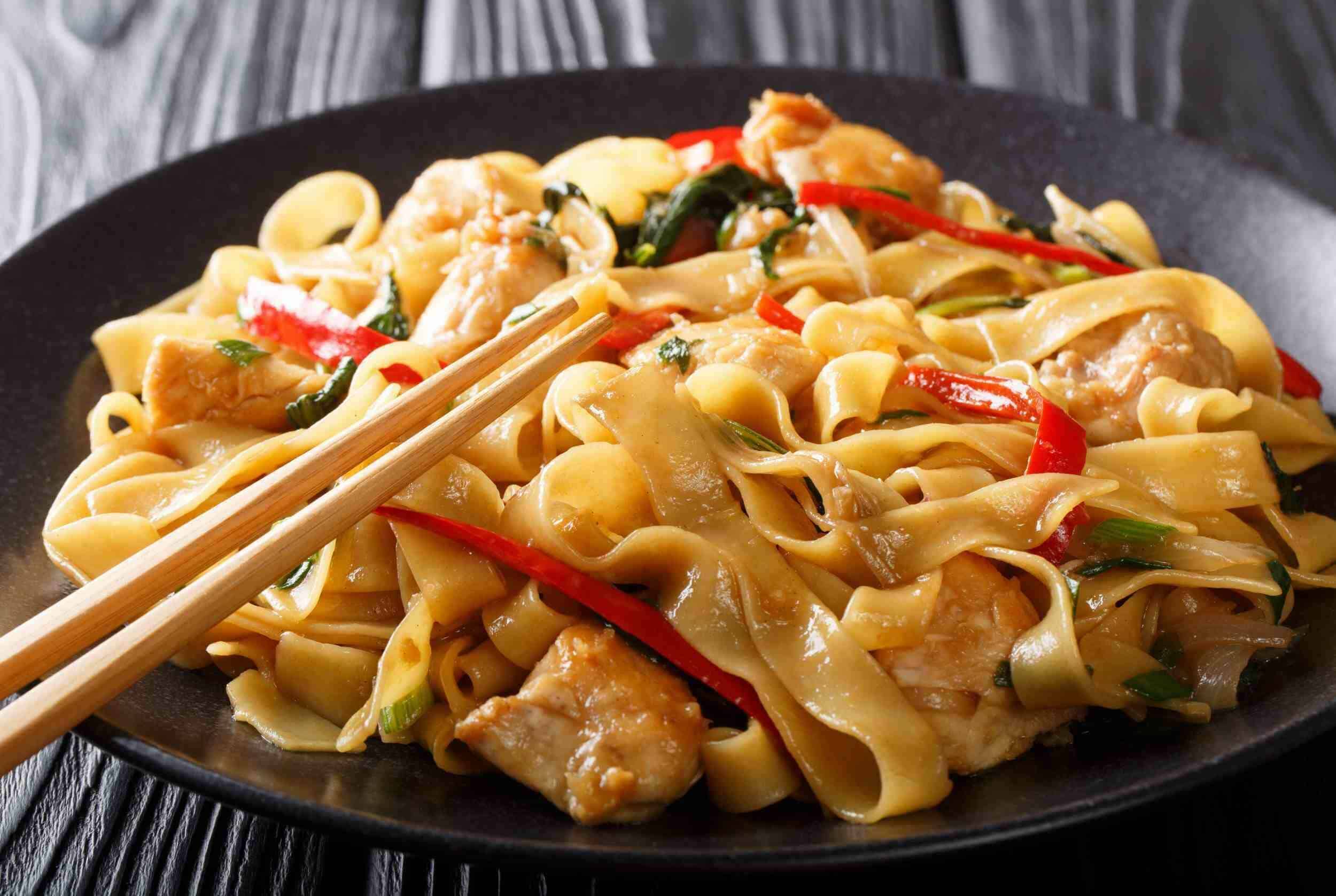 Stir-fried chicken and vegetables with wide egg noodles on a black plate, garnished with red bell peppers and green onions, served on a rustic wooden table.