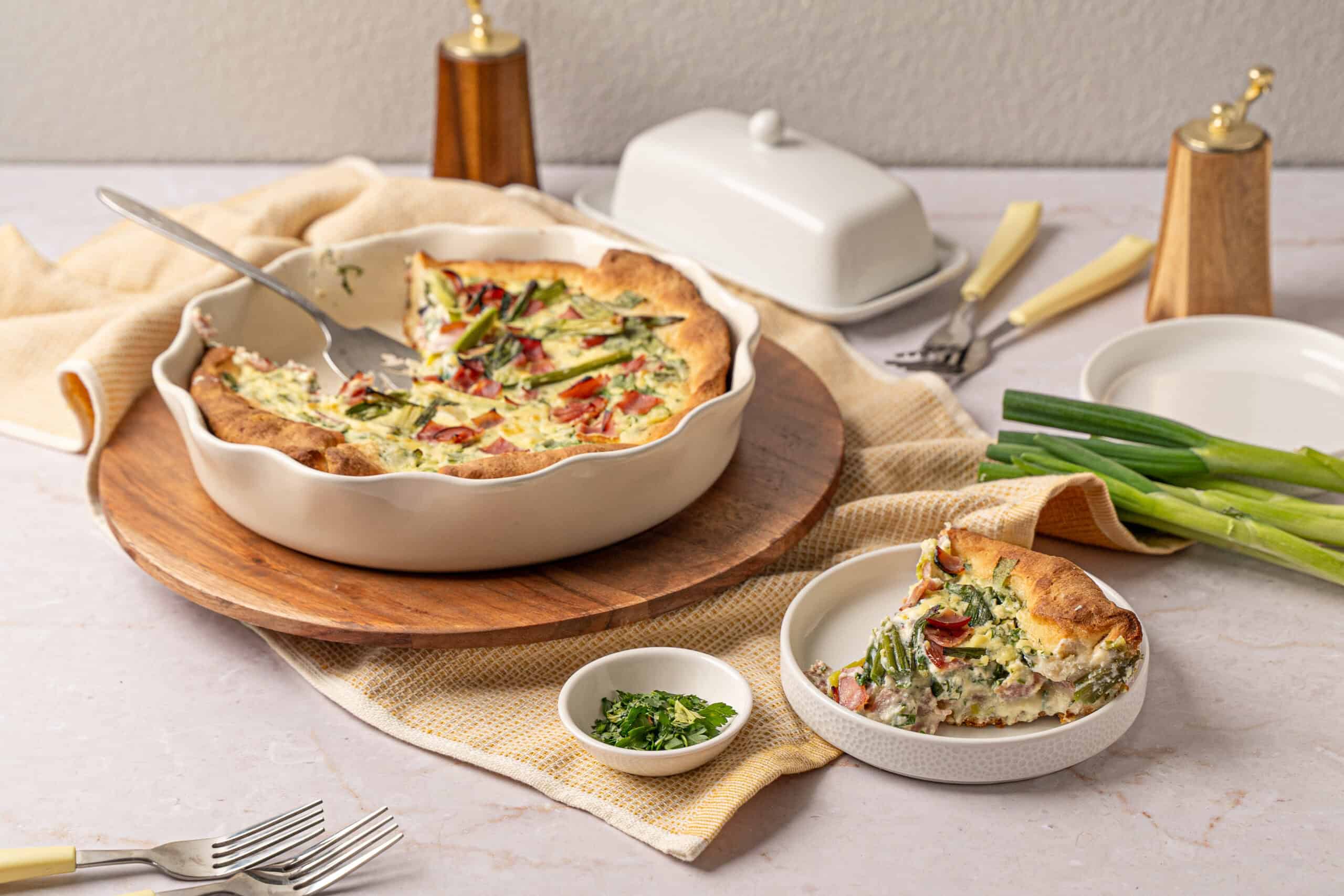 Fluffy vegetable quiche with fresh green onions, served on a white dish with a slice placed on a separate small plate, set on a light-colored table with a rustic napkin and cutlery.