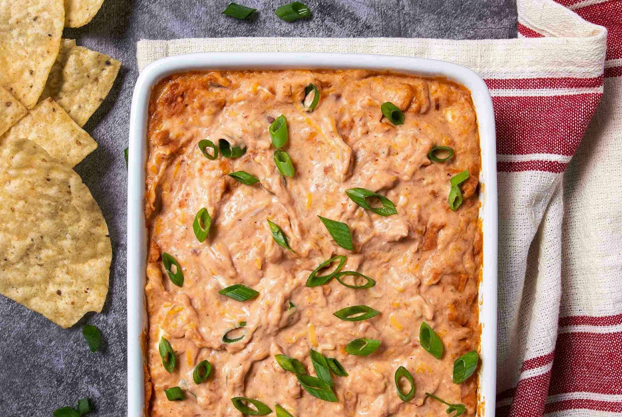 Creamy buffalo chicken dip with melted cheese and green onions in a white serving dish, served with crispy tortilla chips on a rustic surface.