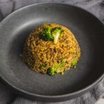 Crispy rice and ground beef stir-fry garnished with broccoli on a black plate, served on a gray cloth, close-up of Asian-style fried rice dish, flavorful and well-seasoned, perfect for dinner or lunch.