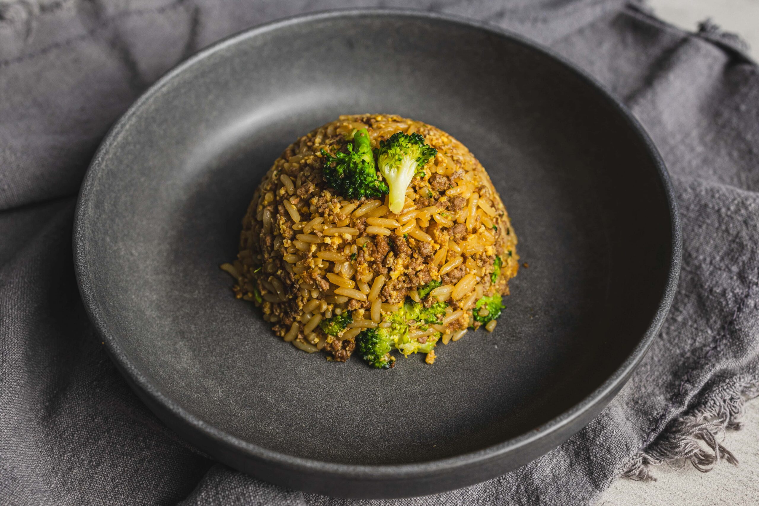 Crispy rice and ground beef stir-fry garnished with broccoli on a black plate, served on a gray cloth, close-up of Asian-style fried rice dish, flavorful and well-seasoned, perfect for dinner or lunch.
