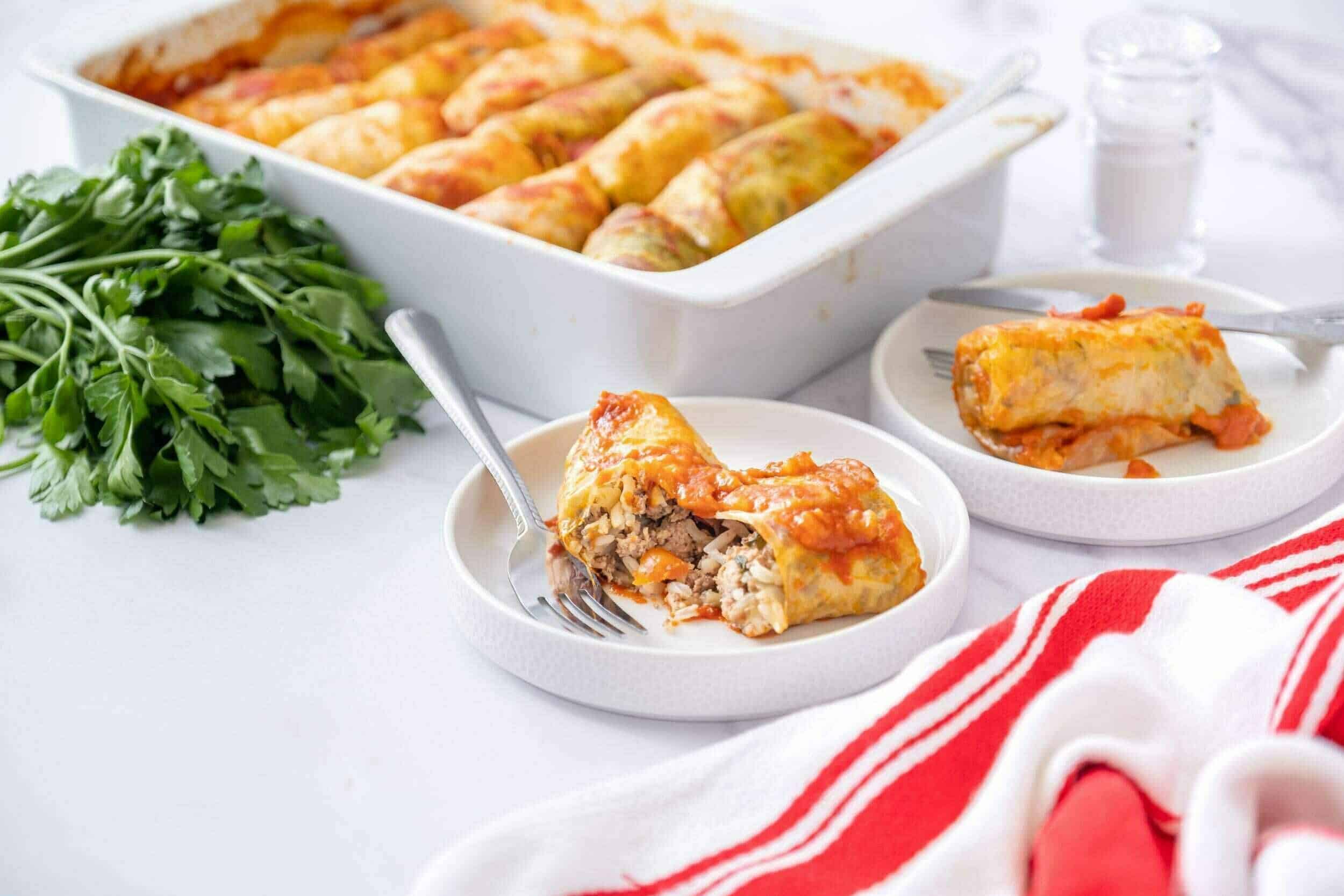 Ground beef and vegetable stuffed cabbage rolls baked in tomato sauce on a white table with fresh parsley, featuring a sliced cabbage roll on a white plate, ideal for hearty, traditional recipes.