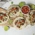 Fresh homemade beef tacos with lettuce, tomato salsa, and avocado slices on a wooden serving board, surrounded by lime wedges and garnished with shredded cheese. Perfect for Mexican cuisine lovers.