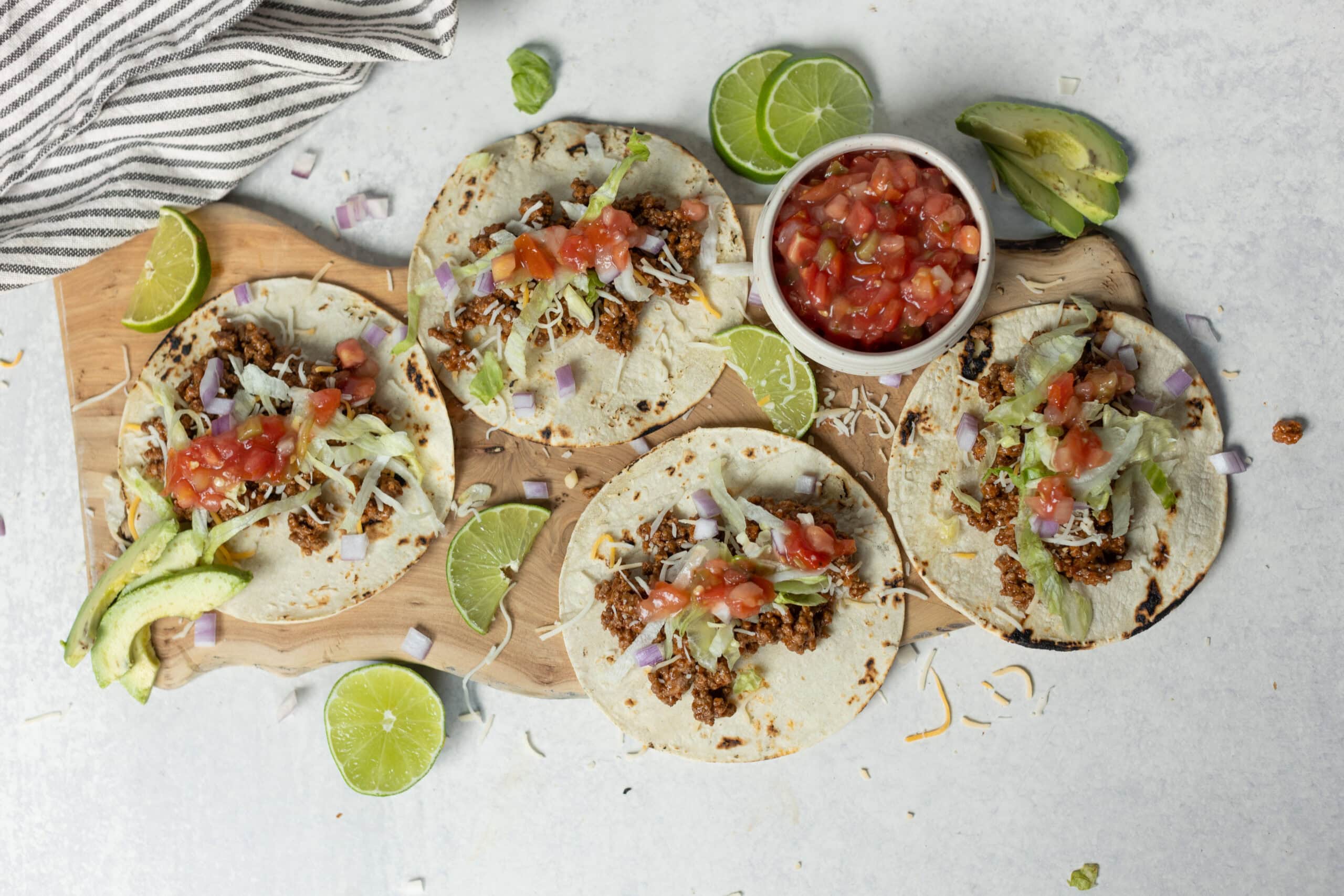 Fresh homemade beef tacos with lettuce, tomato salsa, and avocado slices on a wooden serving board, surrounded by lime wedges and garnished with shredded cheese. Perfect for Mexican cuisine lovers.