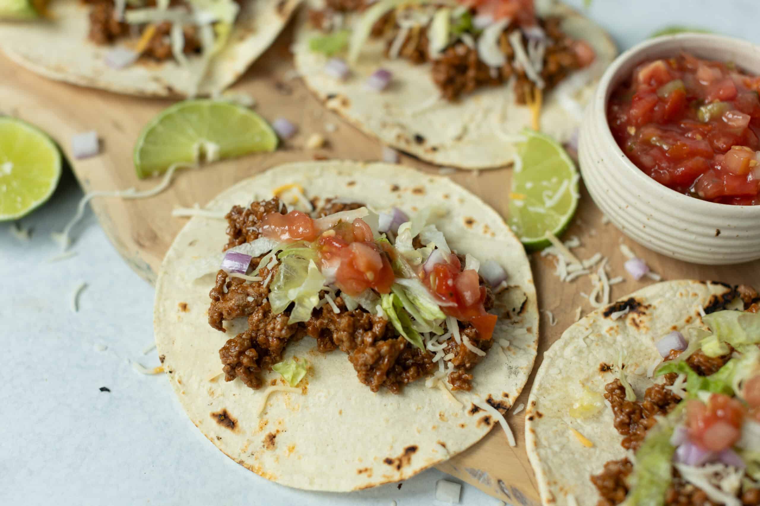 Crunchy beef tacos with fresh lettuce, tomato salsa, shredded cheese, and lime wedges for homemade Mexican-style tacos. Perfect for easy, delicious taco night.