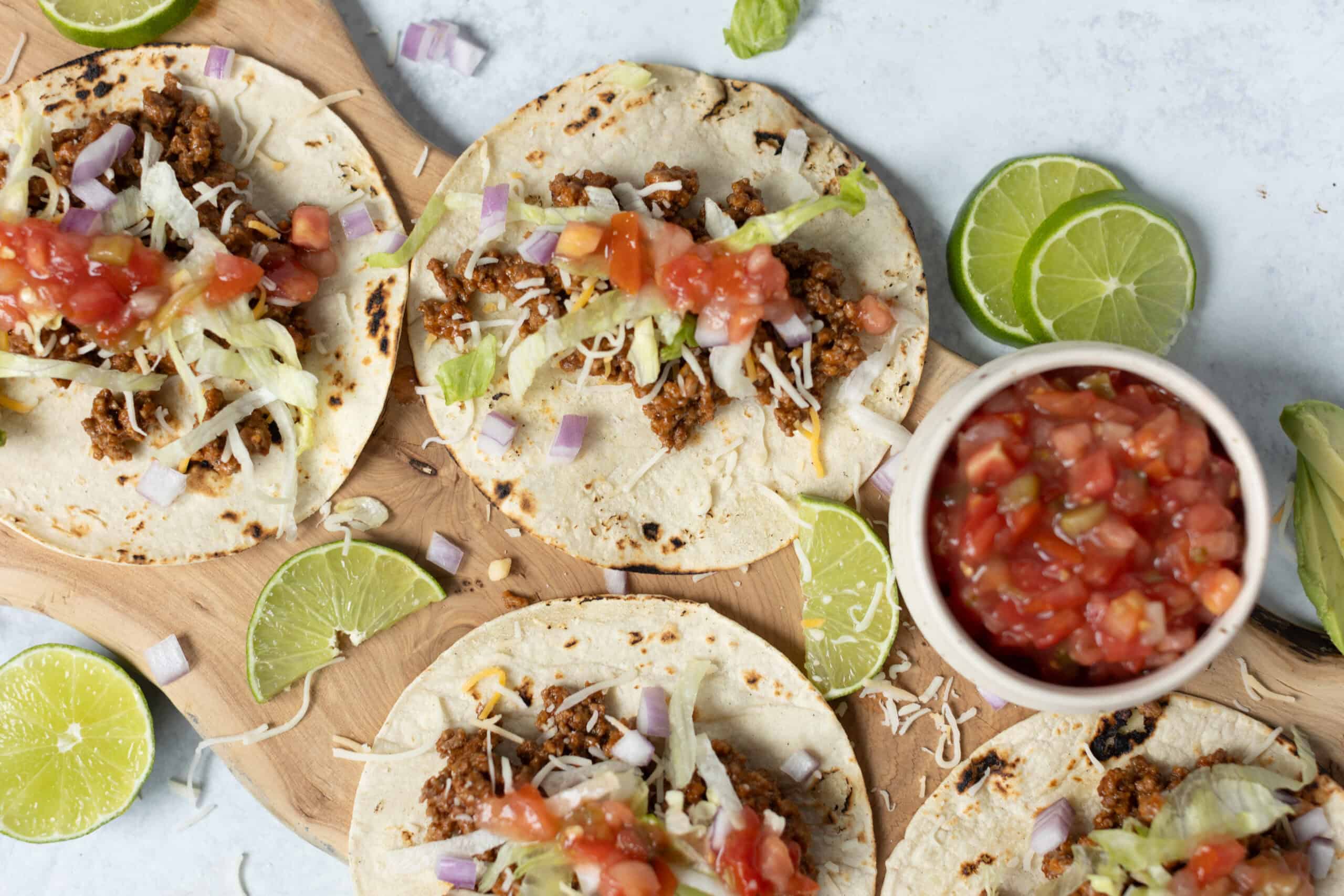 Savory beef tacos with shredded cheese lettuce and tomato in soft tortillas on wooden board with lime wedges and salsa, Mexican street food, easy taco recipe, Tex-Mex cuisine, healthy taco meal.
