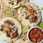 Fresh beef tacos with lettuce, tomato, shredded cheese, diced onions, and lime wedges on a wooden board, served with a side of chunky salsa. Perfect for a tasty Mexican meal or taco night.
