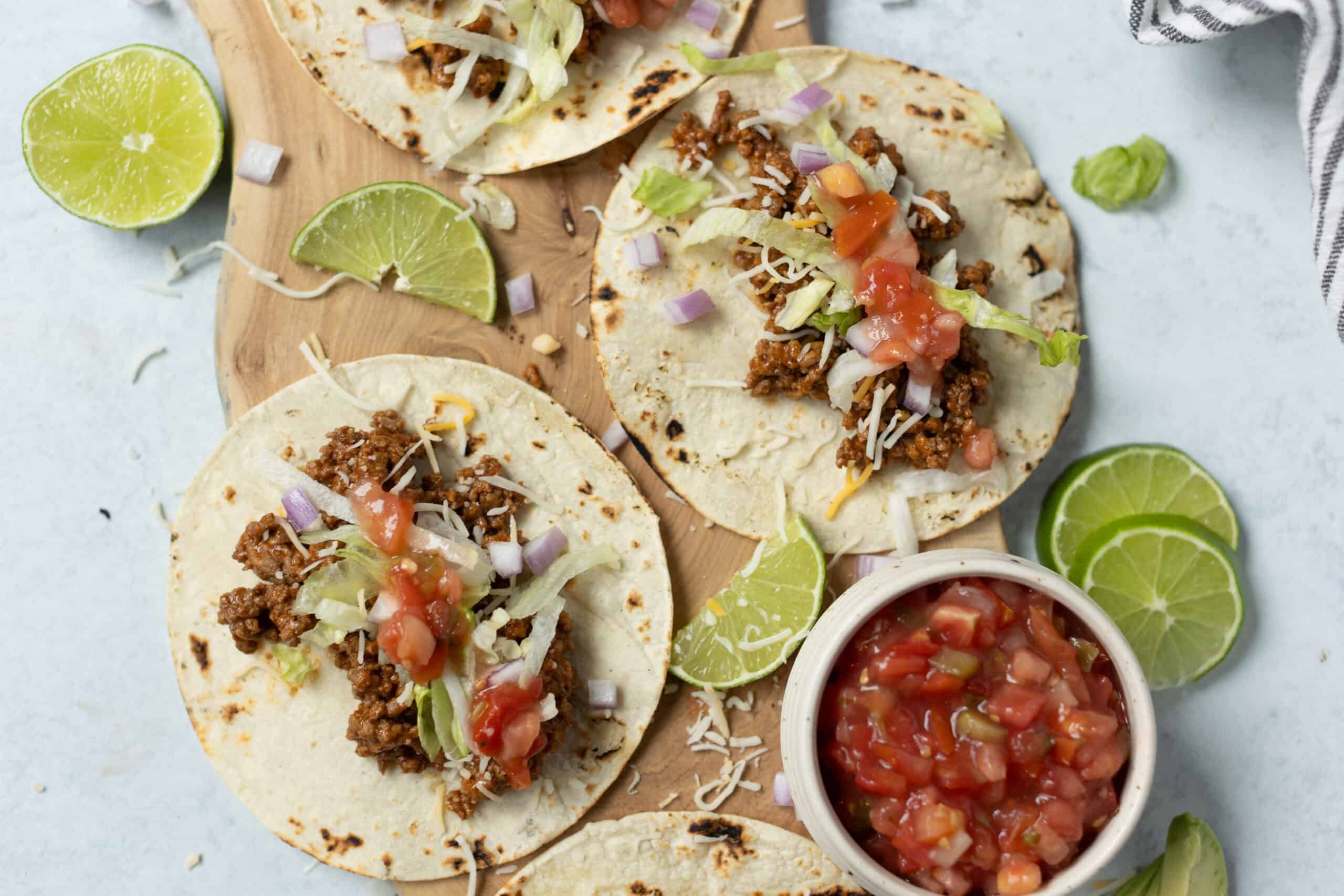 Fresh beef tacos with lettuce, tomato, shredded cheese, diced onions, and lime wedges on a wooden board, served with a side of chunky salsa. Perfect for a tasty Mexican meal or taco night.