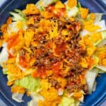 Fresh taco salad with seasoned ground beef, shredded cheese, crispy tortilla chips, and fresh vegetables, topped with tangy salsa, served in a vibrant blue bowl. Perfect for easy, quick meals or gatherings.