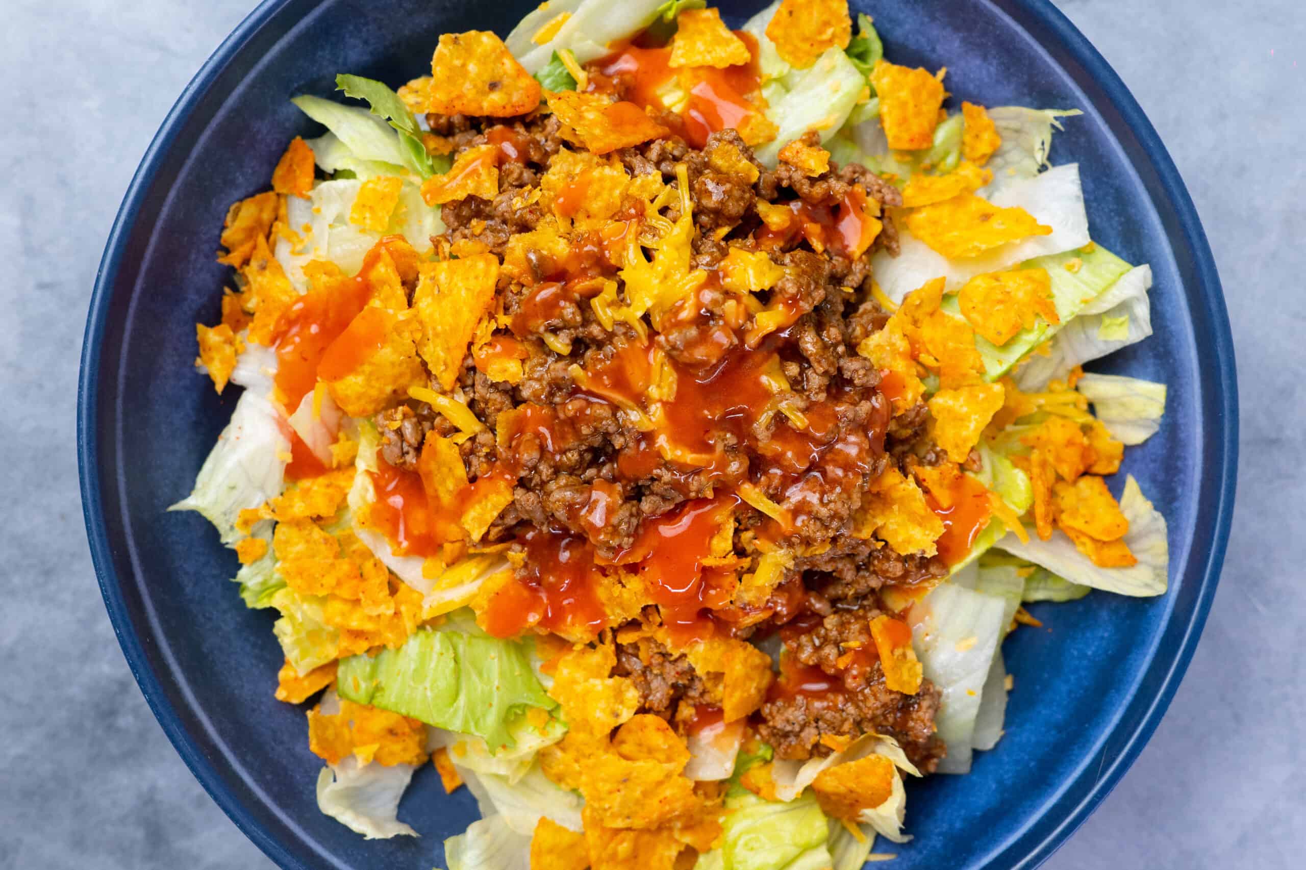 Fresh taco salad with seasoned ground beef, shredded cheese, crispy tortilla chips, and fresh vegetables, topped with tangy salsa, served in a vibrant blue bowl. Perfect for easy, quick meals or gatherings.