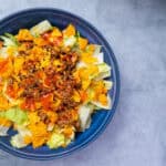Savory taco salad with seasoned ground beef, crispy nacho chips, fresh lettuce, diced tomatoes, shredded cheese, and zesty salsa on a dark blue plate.