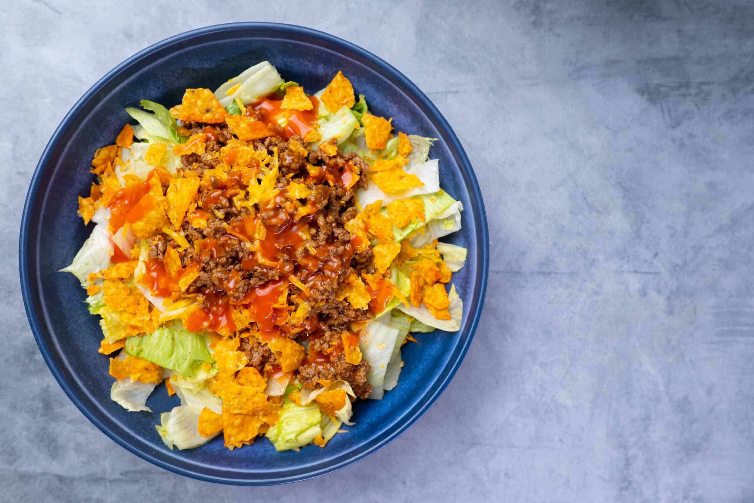 Savory taco salad with seasoned ground beef, crispy nacho chips, fresh lettuce, diced tomatoes, shredded cheese, and zesty salsa on a dark blue plate.