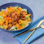 Savory taco salad in a blue bowl with ground beef, shredded cheese, lettuce, and crunchy taco toppings on a gray surface with a blue napkin and gold fork and knife.