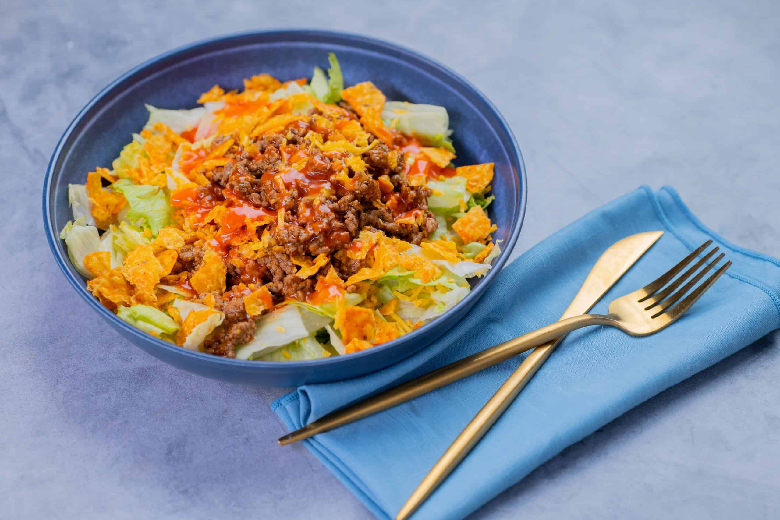 Savory taco salad in a blue bowl with ground beef, shredded cheese, lettuce, and crunchy taco toppings on a gray surface with a blue napkin and gold fork and knife.