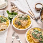 Creamy chicken with spinach and mushroom sauce served over cooked rotini pasta on white bowls, garnished with fresh basil, on a light-colored table with cooking ingredients and utensils.