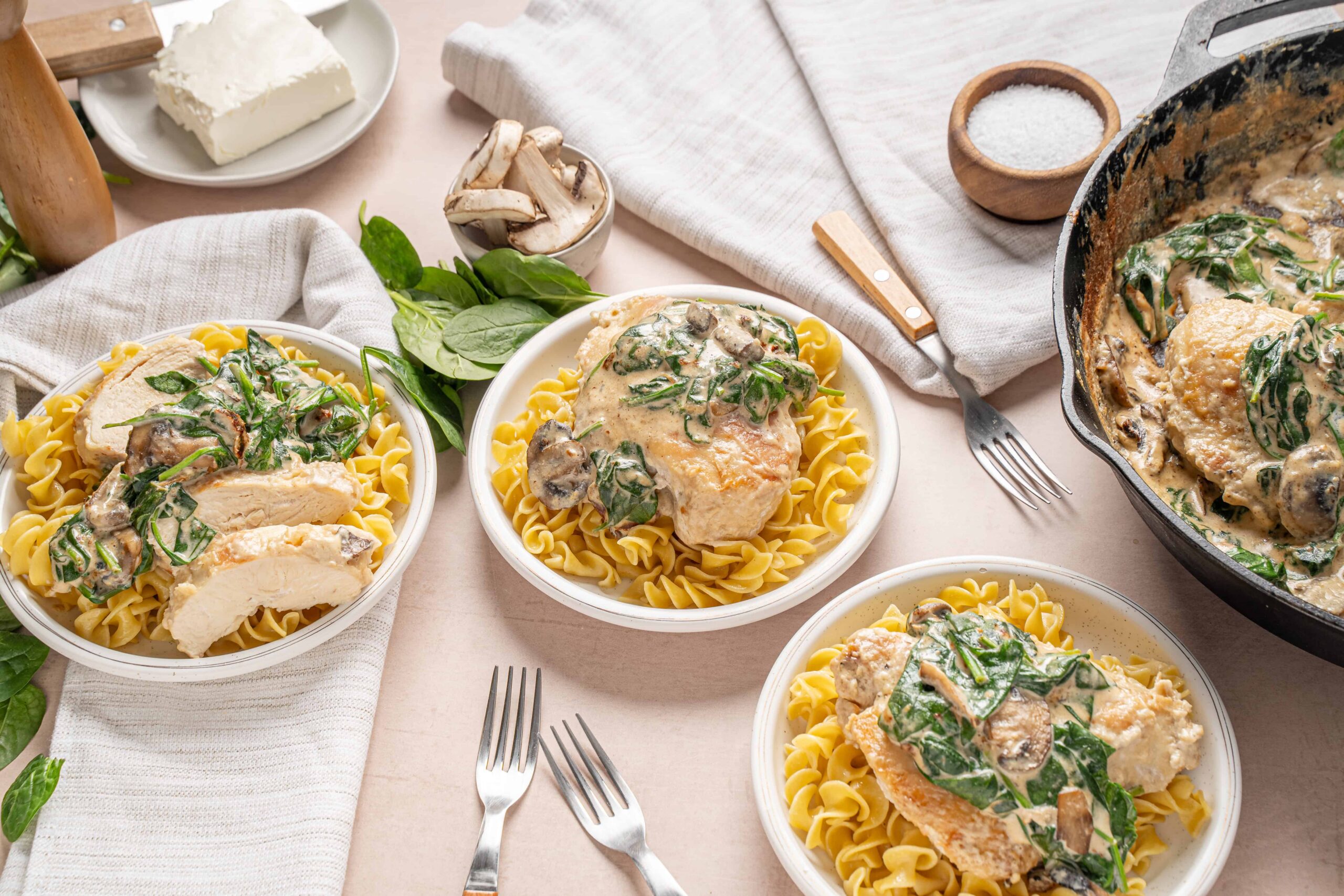 Creamy chicken with spinach and mushroom sauce served over cooked rotini pasta on white bowls, garnished with fresh basil, on a light-colored table with cooking ingredients and utensils.