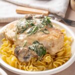 Creamy chicken with mushrooms and spinach served over fusilli pasta in a white dish, featuring tender chicken breast topped with mushroom and spinach cream sauce.