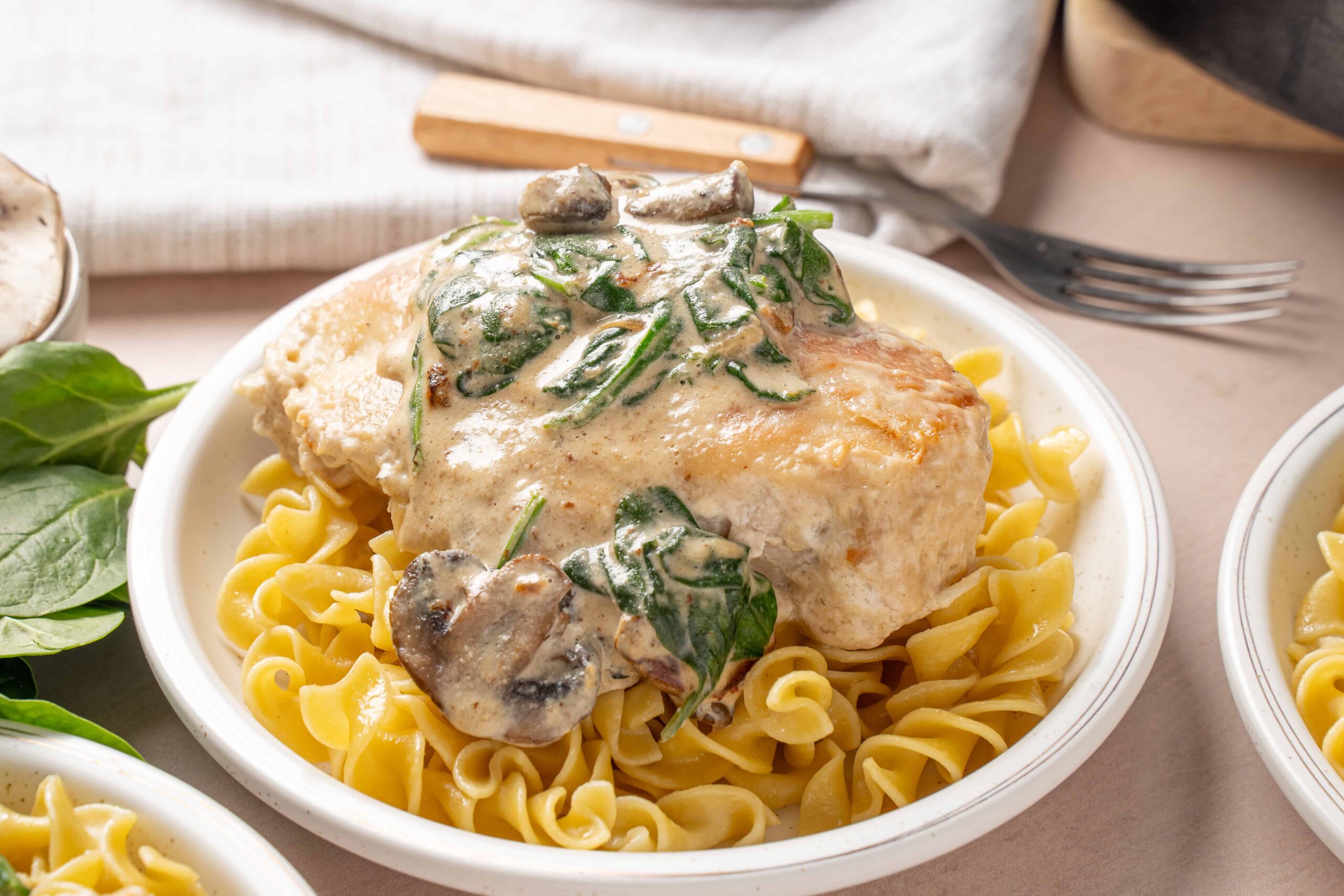 Creamy chicken with mushrooms and spinach served over fusilli pasta in a white dish, featuring tender chicken breast topped with mushroom and spinach cream sauce.
