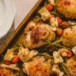 Juicy roasted chicken thighs with cherry tomatoes, grilled asparagus, and rustic bread in a baking dish on a beige surface.