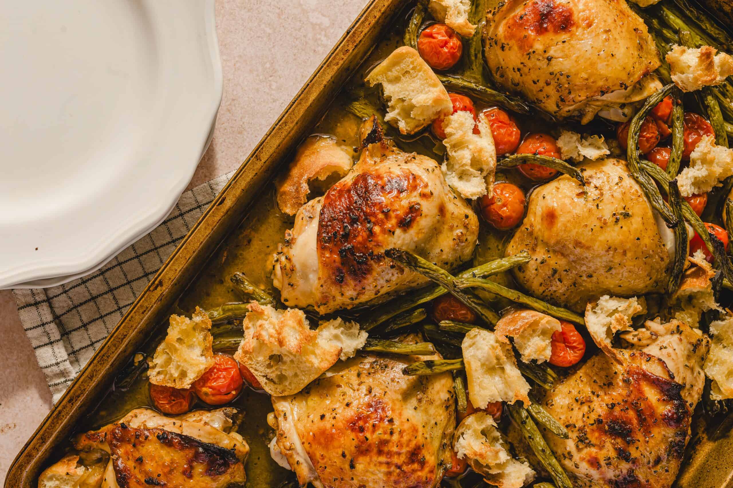Juicy roasted chicken thighs with cherry tomatoes, grilled asparagus, and rustic bread in a baking dish on a beige surface.