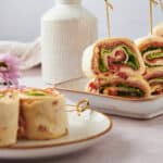Freshly made veggie and ham pinwheels with crisp lettuce, served on a white platter, perfect for appetizers or snacks, displayed with a soft pink flower and a woven ceramic bottle in the background.