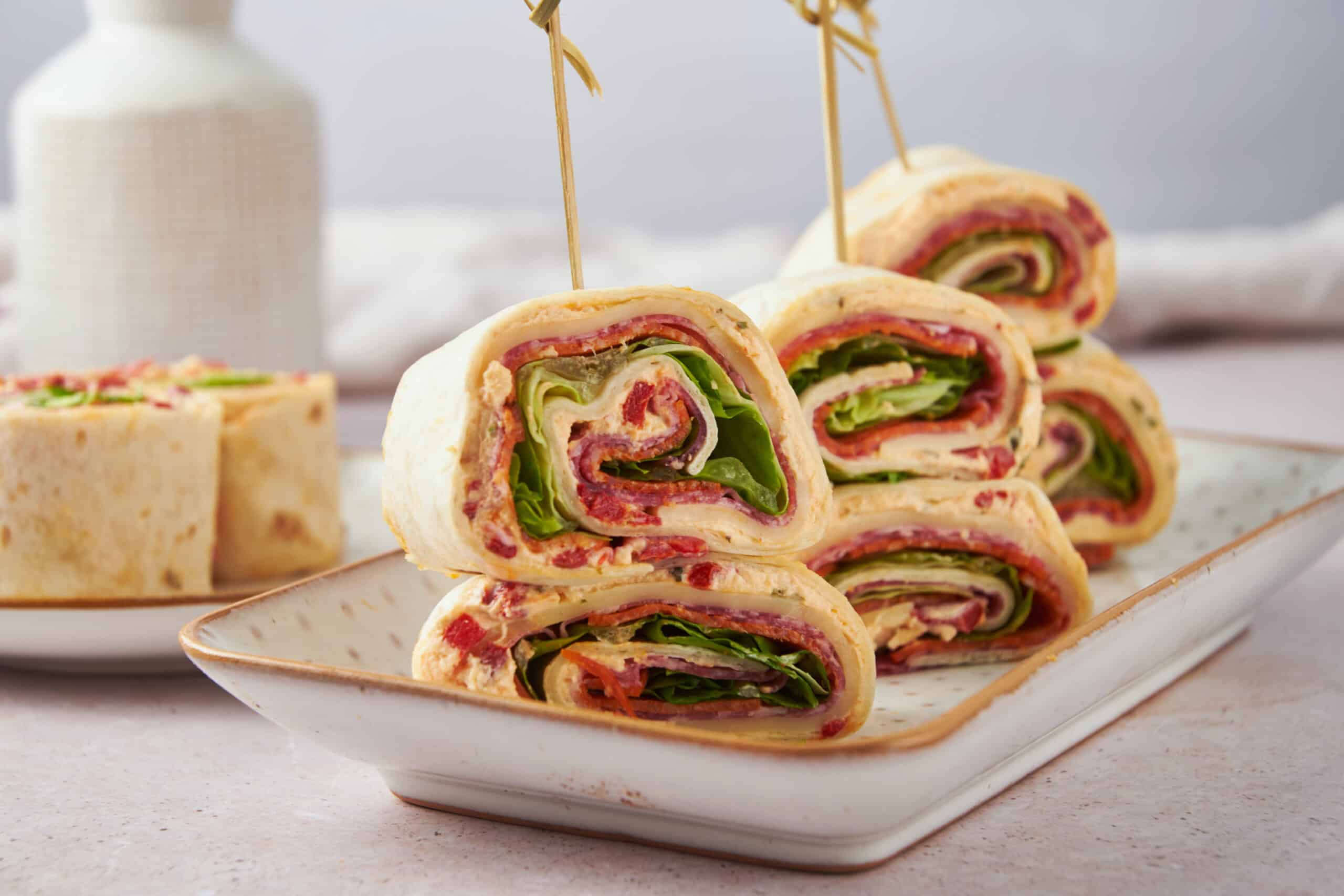 Fresh deli turkey and ham wrap roll-ups with lettuce, tomato, and cheese, sliced into bite-sized pieces for easy snacking or party finger food, served on a stylish white platter.