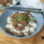 Minced beef over steamed white rice garnished with sliced green onions in a gray bowl, served on a wooden table with chopsticks for an Asian-inspired meal.