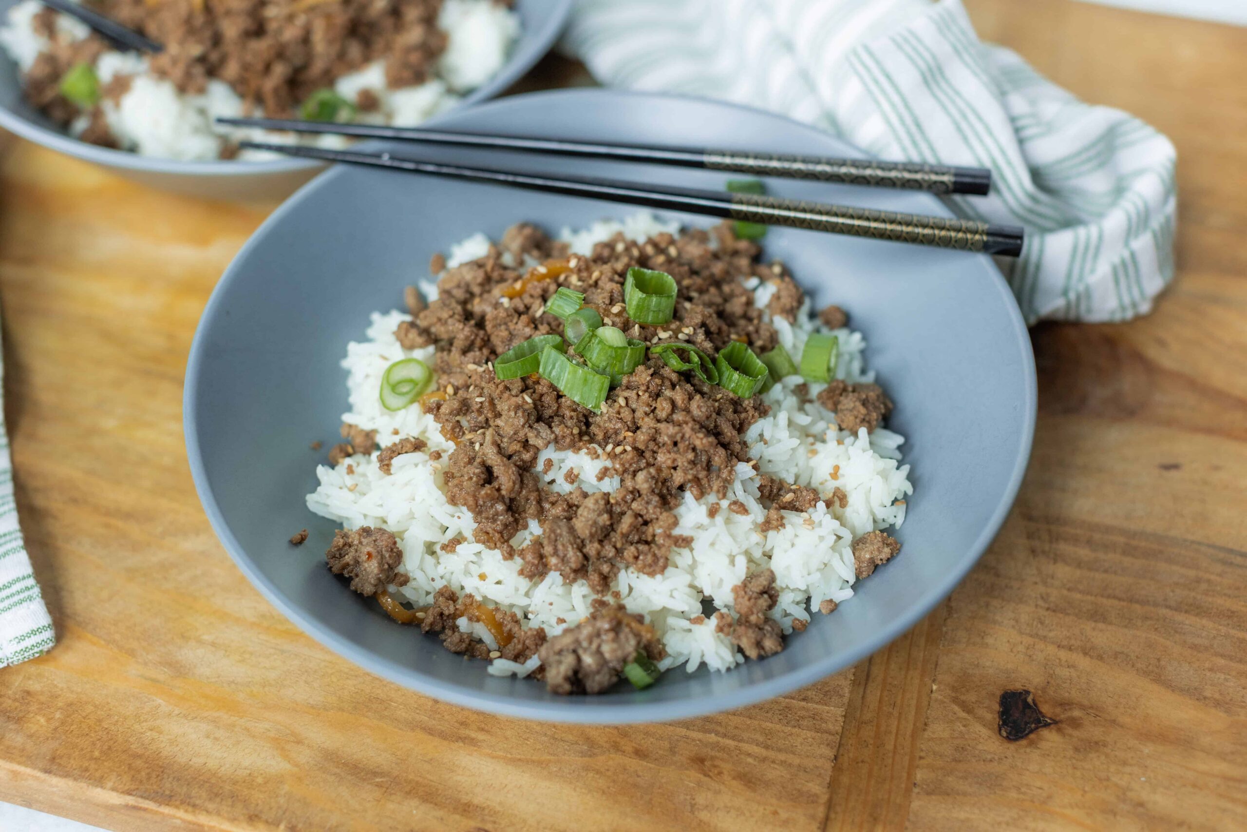 Minced beef over steamed white rice garnished with sliced green onions in a gray bowl, served on a wooden table with chopsticks for an Asian-inspired meal.