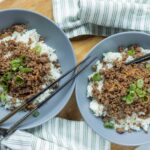 Seasoned ground beef over fluffy white rice garnished with chopped green onions in gray bowls, served on a wooden table with chopsticks and a green-striped napkin, perfect for Asian-inspired meal recipes.