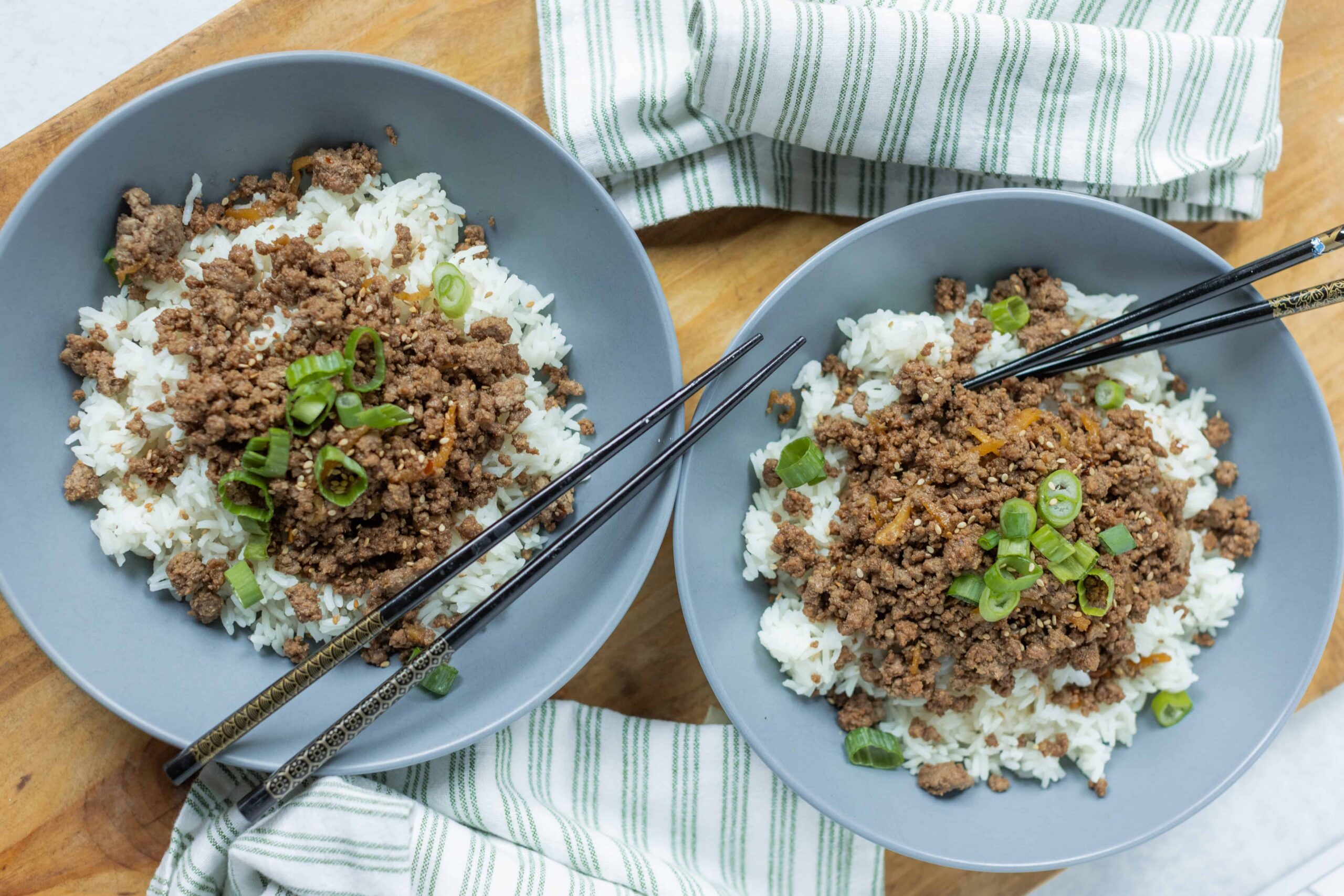 Seasoned ground beef over fluffy white rice garnished with chopped green onions in gray bowls, served on a wooden table with chopsticks and a green-striped napkin, perfect for Asian-inspired meal recipes.