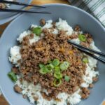 Minced beef stir-fry served over steamed white rice with chopped green onions on top, on a gray plate with black chopsticks, showcasing a delicious Asian-inspired meal for dinner or lunch.