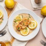 Juicy lemon herb chicken breast served with creamy lemon pasta on white plates, garnished with fresh parsley and lemon slices, perfect for easy summer dinner or lunch.