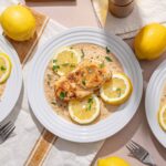Lemon chicken with creamy sauce garnished with fresh lemon slices and chopped parsley on white plates, served with whole lemons on a light-colored table for a fresh, flavorful meal.