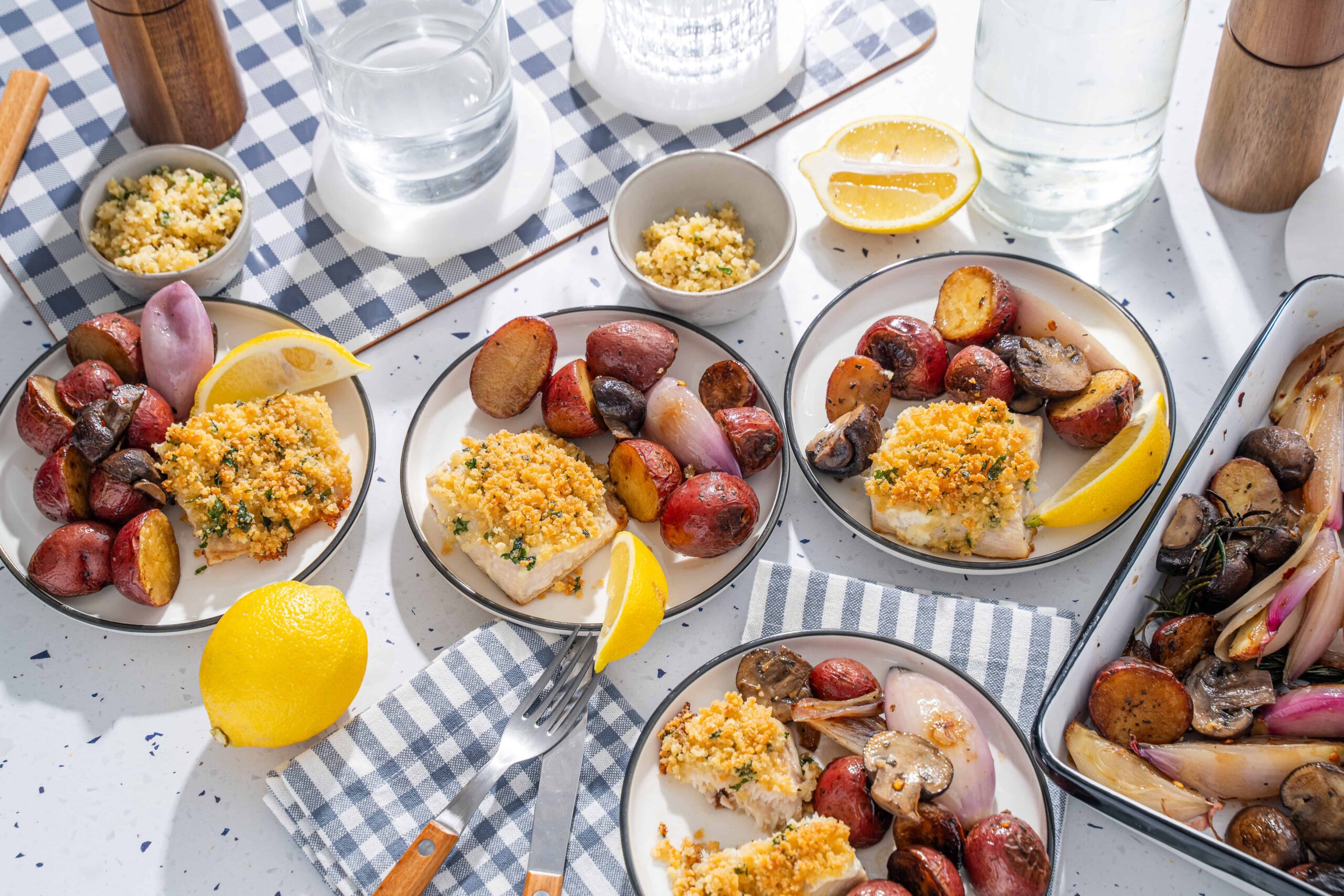 Succulent baked fish fillets topped with golden breadcrumbs, served with roasted red potatoes, onions, and mushrooms on a bright white speckled table, garnished with lemon wedges for a flavorful seafood meal.