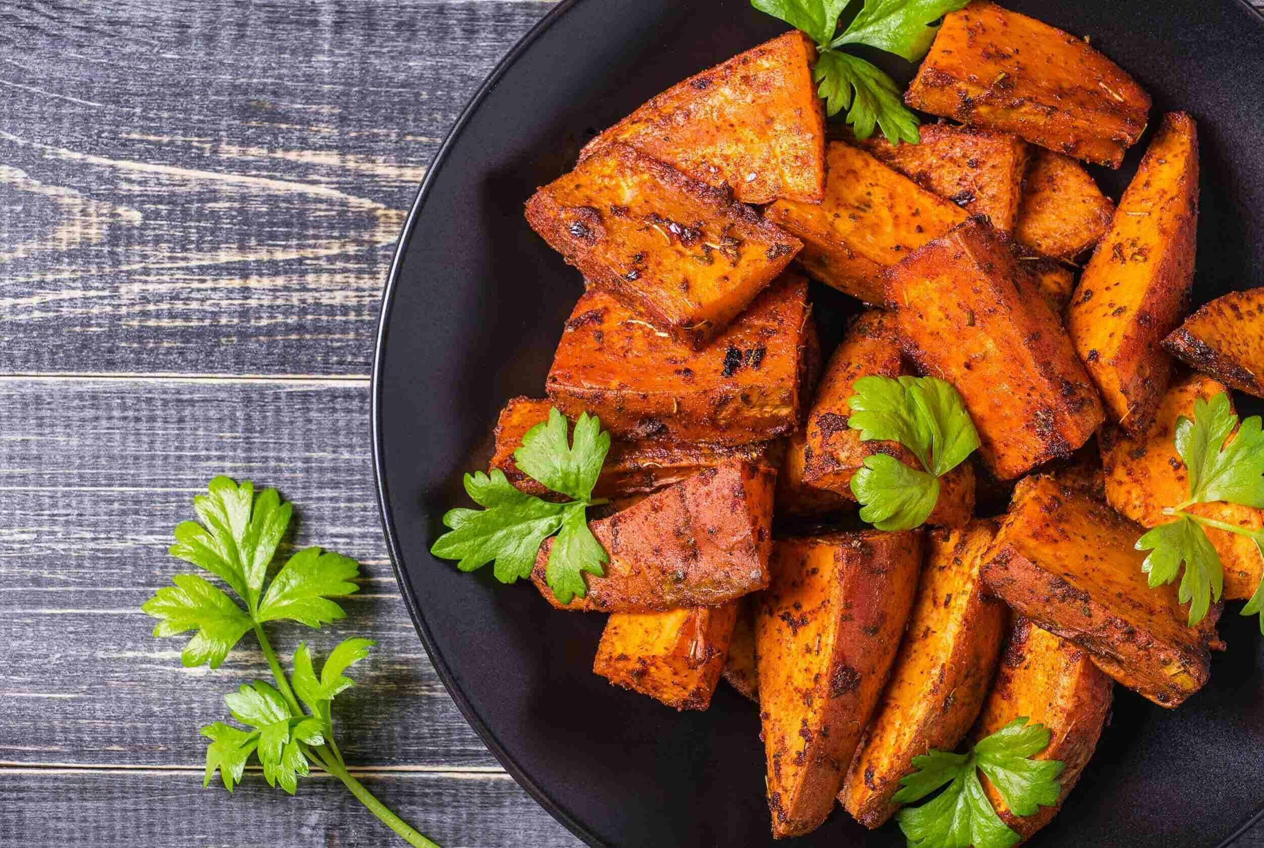 Baked sweet potato wedges garnished with fresh parsley on a black plate. Perfect for healthy snacks or side dishes.