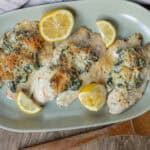 Baked chicken breasts topped with spinach, cheese, and shredded carrots served with lemon wedges on a light green platter.