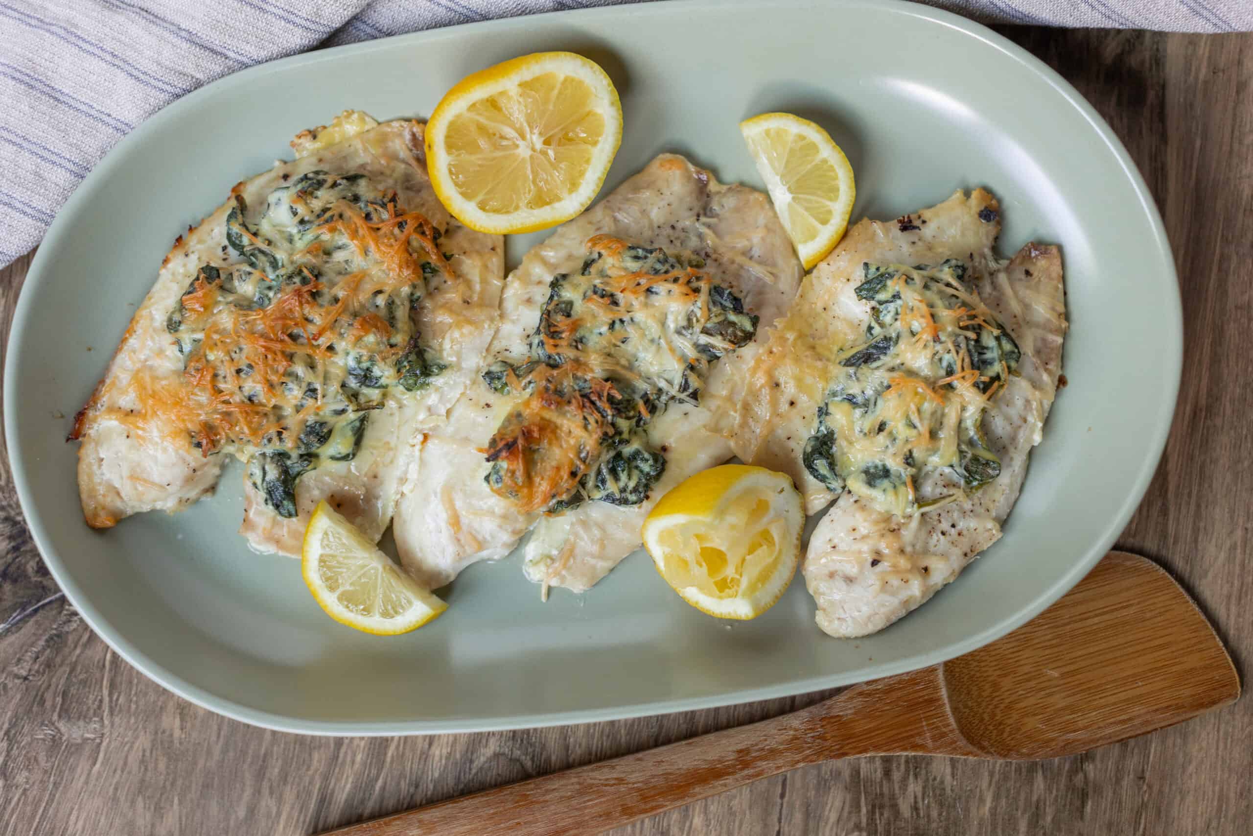 Baked chicken breasts topped with spinach, cheese, and shredded carrots served with lemon wedges on a light green platter.