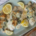 Baked cod fillets topped with spinach, cheese, and herbs, served with lemon wedges on a green platter. Perfect for healthy, delicious seafood dinner recipes.