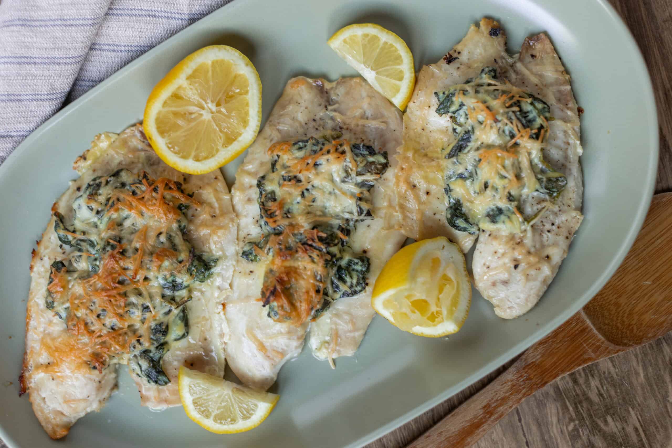 Baked cod fillets topped with spinach, cheese, and herbs, served with lemon wedges on a green platter. Perfect for healthy, delicious seafood dinner recipes.