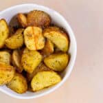 Roasted garlic herb potatoes in a white bowl on a beige background. Perfect side dish for dinner recipes and healthy eating. Easy baked potato wedges with seasoning and herbs.
