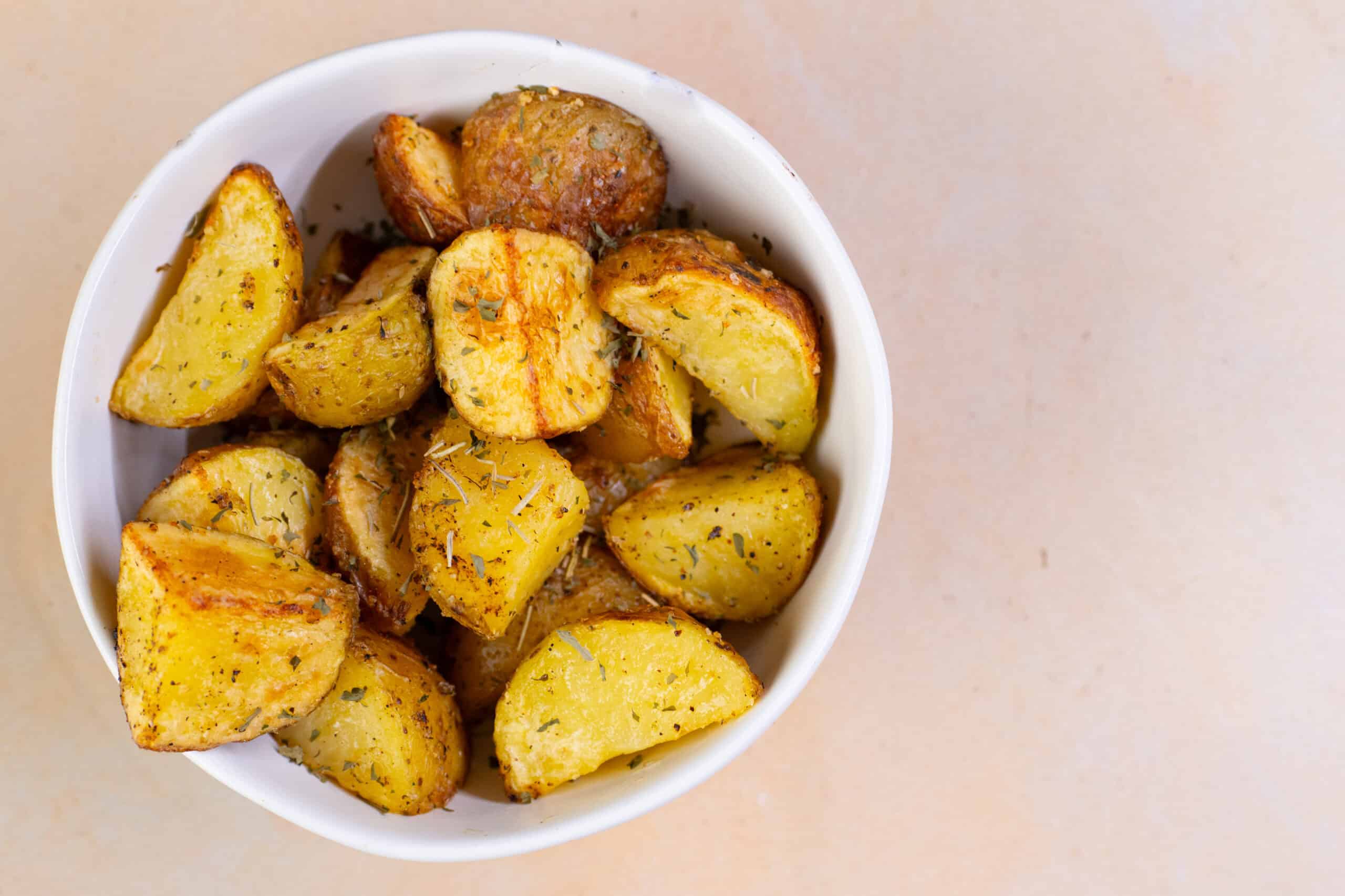 Roasted garlic herb potatoes in a white bowl on a beige background. Perfect side dish for dinner recipes and healthy eating. Easy baked potato wedges with seasoning and herbs.