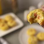 Soft, freshly baked breakfast muffin with visible bits of tomato, green pepper, and cheese, held by a hand, with plates of similar muffins and a yellow drink in the blurred background.