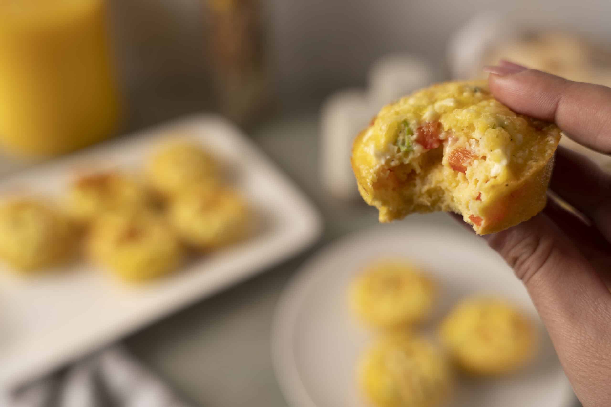 Soft, freshly baked breakfast muffin with visible bits of tomato, green pepper, and cheese, held by a hand, with plates of similar muffins and a yellow drink in the blurred background.