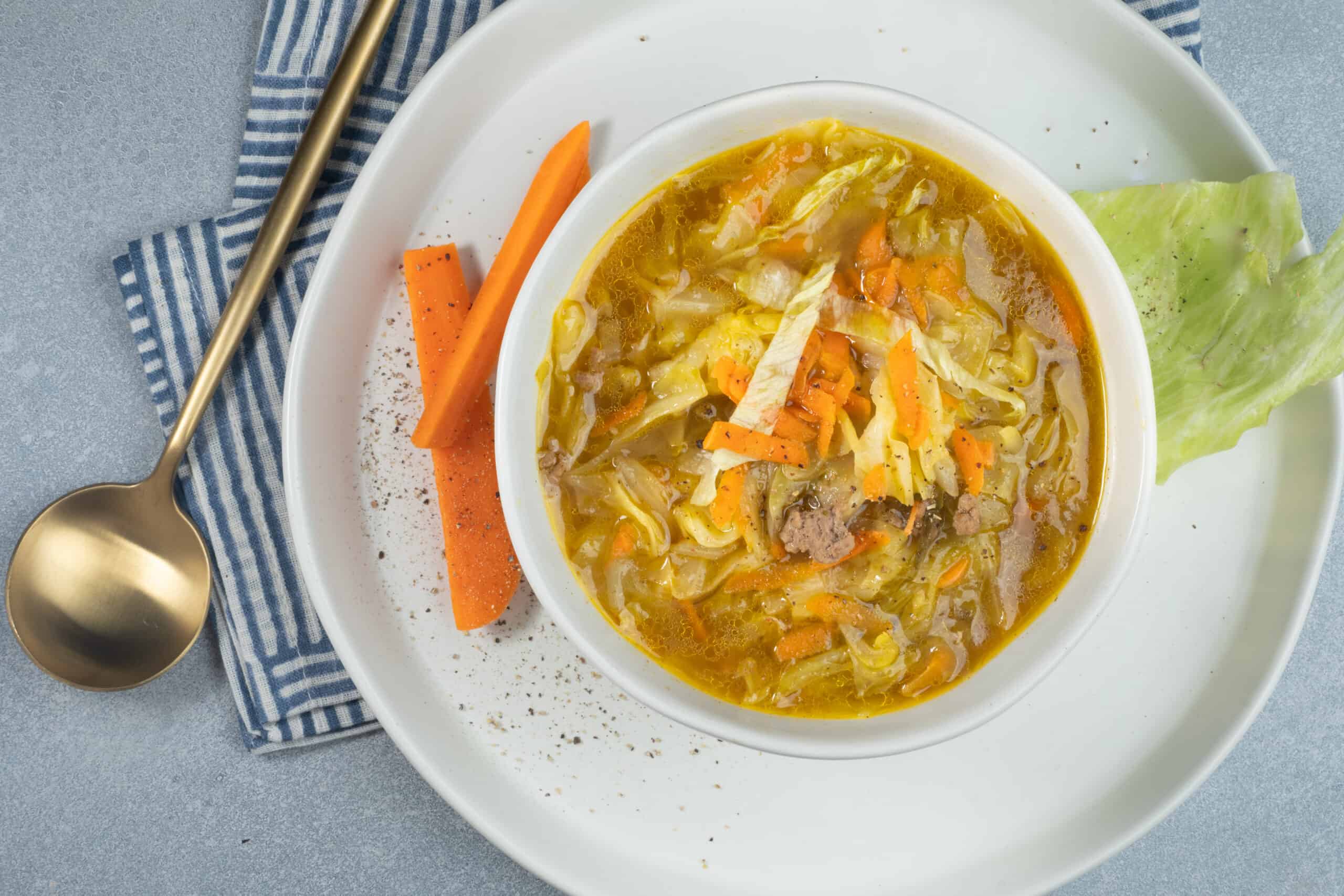 A bowl of homemade vegetable cabbage soup with carrots, ground meat, and shredded cheese, garnished with a lettuce leaf and served on a white plate.