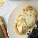 Creamy chicken and mashed potatoes topped with herbs served alongside black rice on a white plate. Perfect comfort food pairing with a glass of white wine.