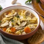Creamy chicken noodle soup with vegetables in a rustic bowl, garnished with fresh herbs, served with crackers and spices for a comforting homemade meal.