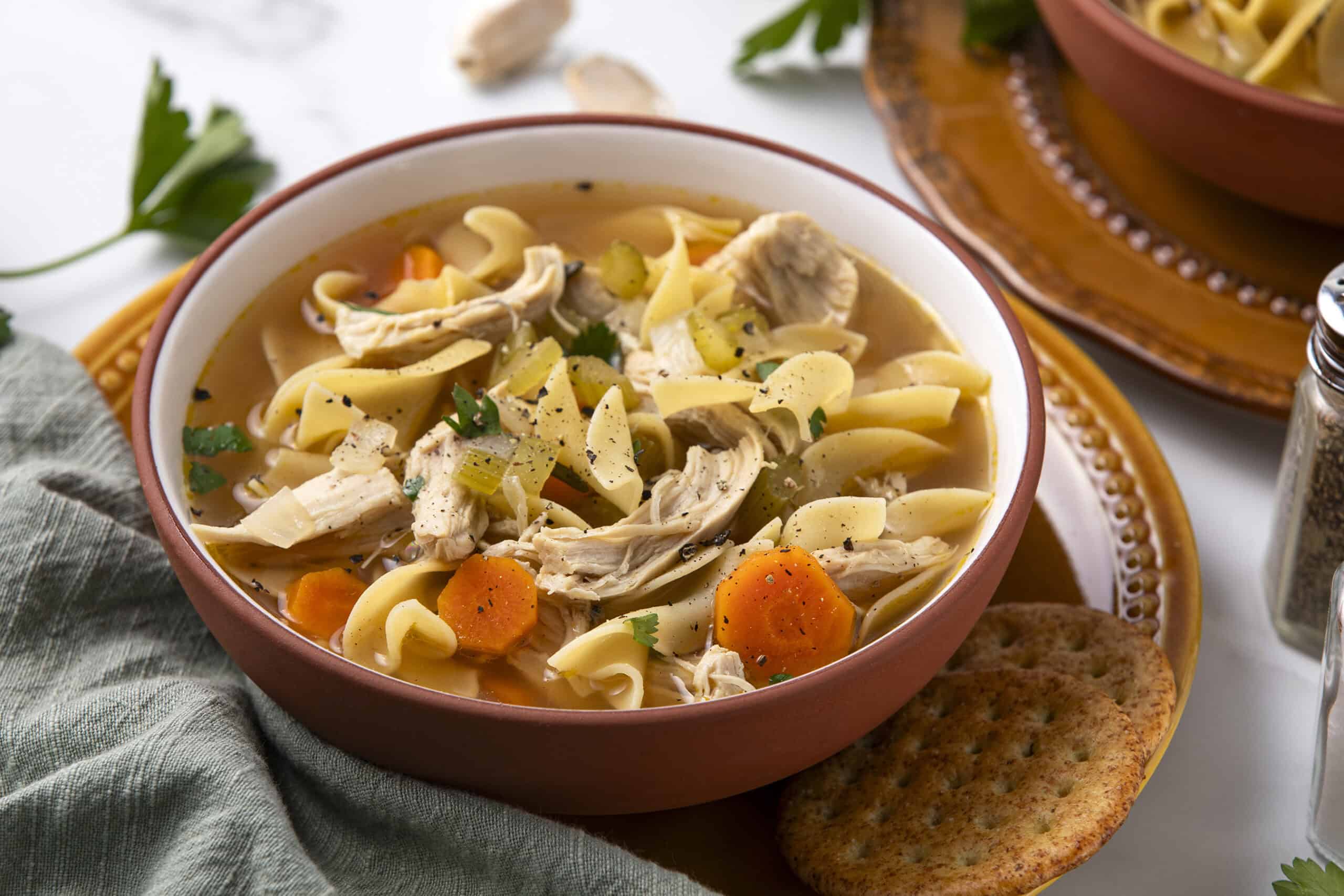 Creamy chicken noodle soup with vegetables in a rustic bowl, garnished with fresh herbs, served with crackers and spices for a comforting homemade meal.